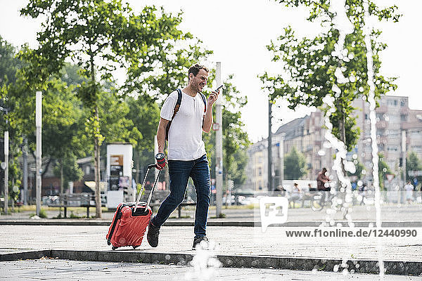 Man with rolling suitcase and cell phone walking at fountain in the city