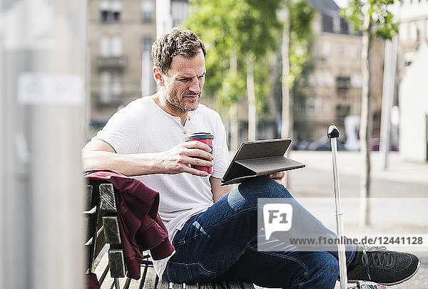 Man with rolling suitcase and takeaway coffee sitting on bench using tablet