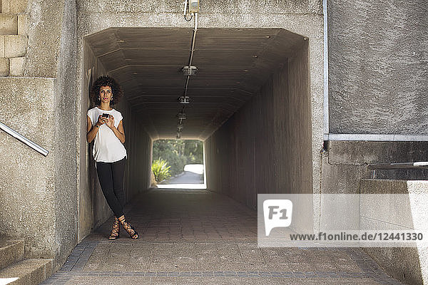 Young woman with smartphone leaning in underpass