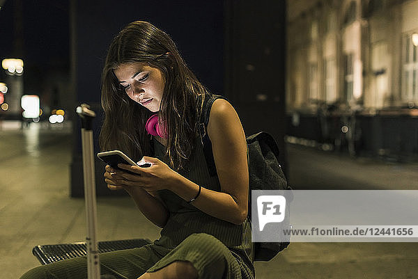Young woman with backpack and baggage using cell phone at night