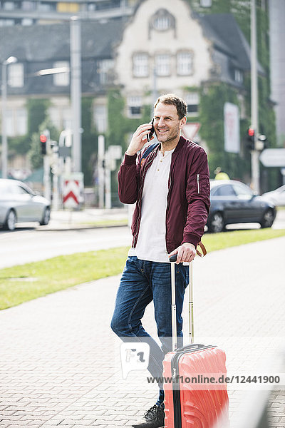 Smiling man with rolling suitcase on cell phone in the city