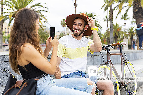 Spain  Barcelona  happy couple sitting on bench taking a smartphone picture