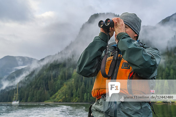 Man looking through a pair of binoculars with his sailboat in the distance  Great Bear Rainforest; Hartley Bay  British Columbia  Canada