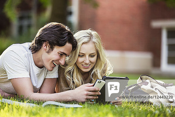 Ein junges Paar studiert im Freien auf dem Rasen des Universitätscampus mit einem Lehrbuch und einem Tablet und überprüft die sozialen Medien auf einem Smartphone,  Edmonton,  Alberta,  Kanada