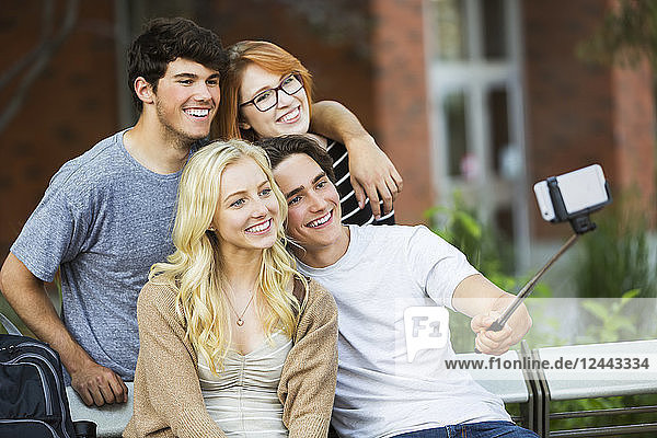 Eine Gruppe von vier Freunden sitzt auf einer Bank und macht ein Selbstporträt mit einem Smartphone und einem Selfie-Stick auf dem Universitätscampus in Edmonton,  Alberta,  Kanada