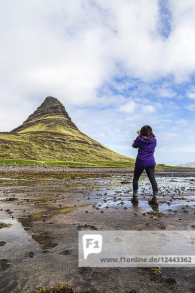 A female hiker stops on the beach at low tide to take a photo with an SLR camera in front of Kirkjufell Mountain in Snaefellsnes peninsula  Western Iceland  Iceland