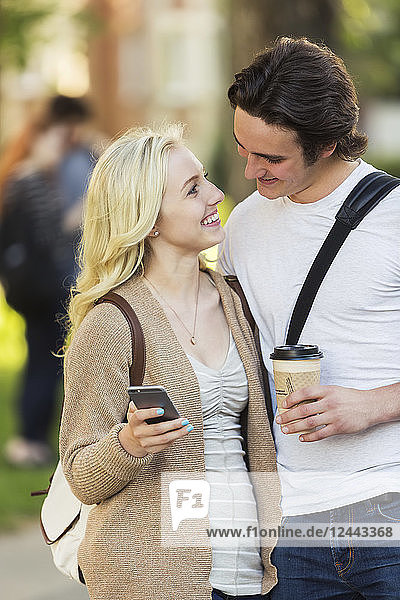 Ein junges Paar,  das sich in die Augen schaut und soziale Medien auf einem Smartphone abruft,  während es durch einen Universitätscampus geht,  Edmonton,  Alberta,  Kanada
