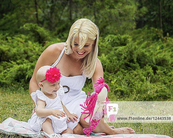 A beautiful young mother enjoying quality time with her cute baby daughter sitting on a blanket and playing with a doll in a city park on a summer day  Edmonton  Alberta  Canada.