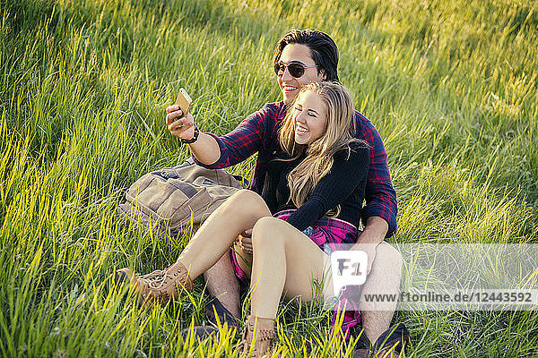 Young couple sit on the grass in a park posing for a self-portrait with their cell phone  Edmonton  Alberta  Canada