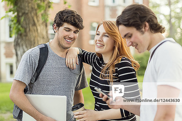 Drei Freunde stehen auf dem Universitätscampus und unterhalten sich mit ihrem Smartphone und Laptop,  Edmonton,  Alberta,  Kanada