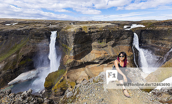 A young asian female hiker poses for a portrait on the edge of a stunning double waterfall valley landscape known as Haifoss  Iceland
