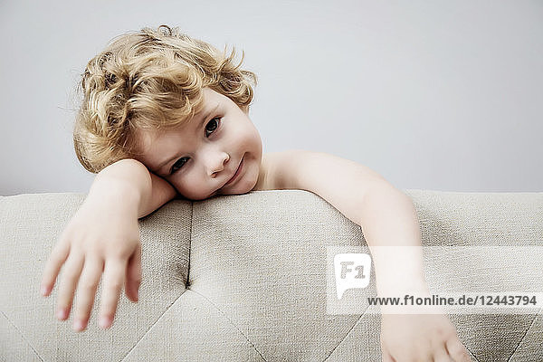 Ein niedlicher kleiner Junge mit langem  lockigem  blondem Haar  der sich an die Rückenlehne einer Couch lehnt und müde aussieht; Langley  British Columbia  Kanada