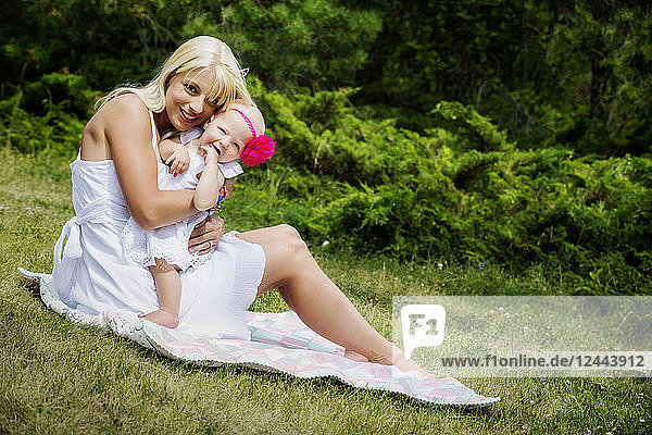 Eine hübsche junge Mutter mit langen blonden Haaren genießt die Zeit mit ihrer süßen kleinen Tochter auf einer Decke im Gras eines Stadtparks an einem Sommertag  Edmonton  Alberta  Kanada