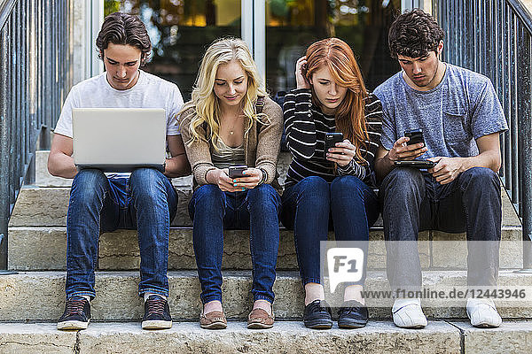 Vier Studenten sitzen in einer Reihe auf einer Stufe und nutzen ihre Technologie auf dem Universitätscampus,  Edmonton,  Alberta,  Kanada