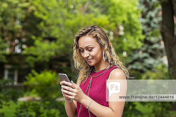 Eine hübsche junge Frau  die draußen auf einem Universitätscampus eine SMS auf ihrem Smartphone schreibt  Edmonton  Alberta  Kanada