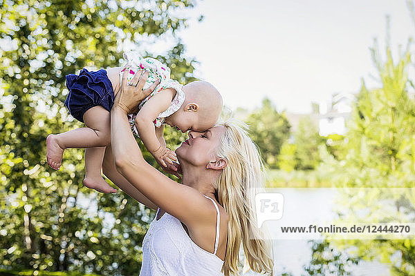 A beautiful young mother with long blonde hair enjoying quality time with her cute baby daughter and tossing her in the air in a city park on a summer day  Edmonton  Alberta  Canada