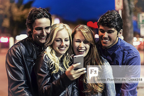 Eine Gruppe von vier Freunden kauert auf einem Bürgersteig zusammen und schaut auf ein Smartphone,  während das Leuchten des Bildschirms ihre Gesichter erhellt,  Edmonton,  Alberta,  Kanada