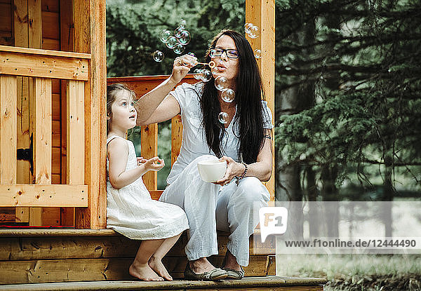 Mother and young daughter blowing bubbles on the steps of a wooden playhouse; Alberta  Canada