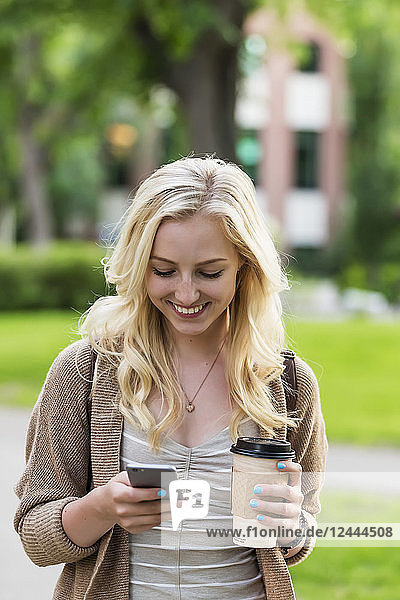 Eine schöne junge Frau mit langen blonden Haaren hält eine Kaffeetasse und schreibt eine SMS auf ihrem Smartphone,  während sie über einen Universitätscampus geht,  Edmonton,  Alberta,  Kanada
