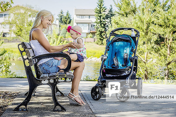 Eine hübsche junge Mutter hält ihre kleine Tochter auf den Knien  während sie sich auf einer Parkbank am See ausruht  nachdem sie an einem warmen sonnigen Tag mit einem Kinderwagen spazieren gegangen ist  Edmonton  Alberta  Kanada