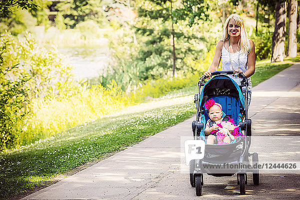 A beautiful young mother taking her baby daughter out for a walk using a stroller in a park with a lake on a warm sunny day  Edmonton  Alberta  Canada