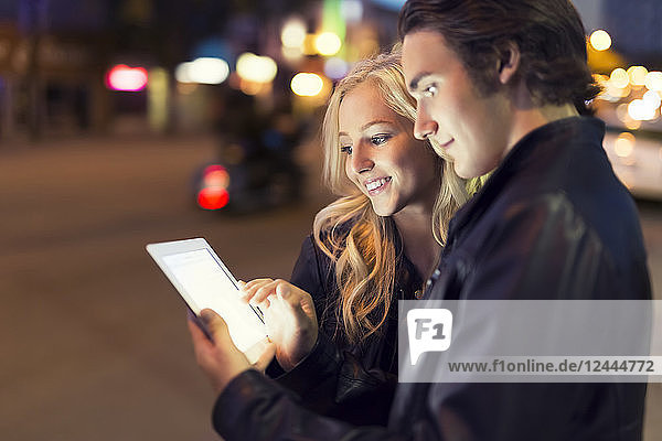 Ein junges Paar benutzt ein Tablet auf einer Straße in der Abenddämmerung  wobei der leuchtende Bildschirm ihre Gesichter erhellt  Edmonton  Alberta  Kanada