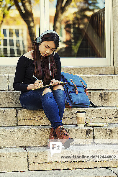 Eine junge chinesische Studentin sitzt auf einer Treppe  hört Musik über Kopfhörer und schreibt in ein Notizbuch  Edmonton  Alberta  Kanada