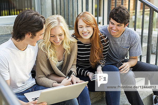 Vier Studenten,  die in einer Reihe auf einer Stufe sitzen,  ihre Technologie auf dem Universitätscampus nutzen und zusammen lachen,  Edmonton,  Alberta,  Kanada