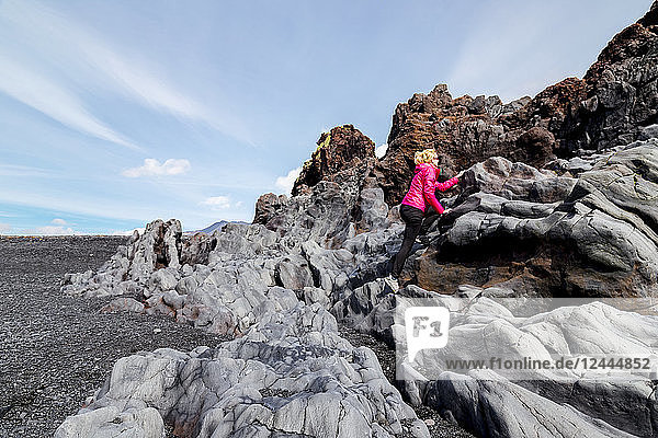 A female hiker climbs a lava rock formation on the black sand beach in Western Iceland,  Iceland