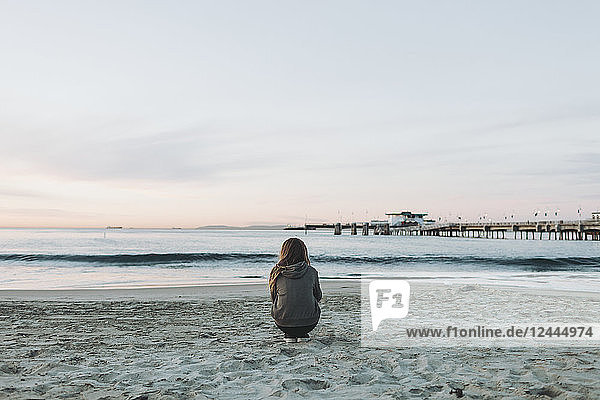 Ein Mädchen hockt am Strand und blickt bei Sonnenuntergang über den Ozean auf den Horizont; Long Beach  Kalifornien  Vereinigte Staaten von Amerika
