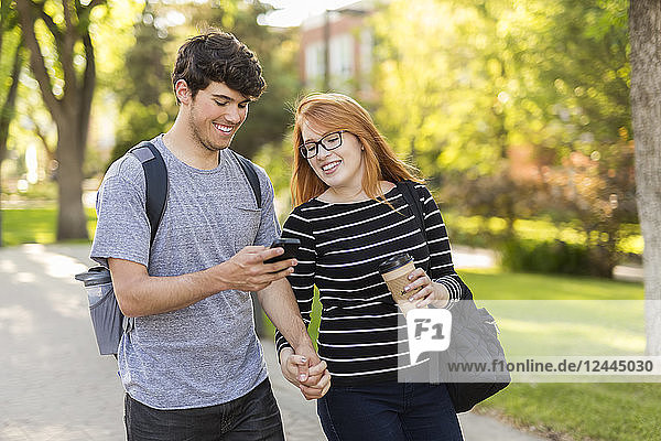 Ein junges Paar  das zusammen spazieren geht und auf dem Universitätsgelände in Edmonton  Alberta  Kanada  soziale Medien auf seinem Smartphone abruft.