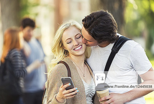 A young man kisses a young woman on the cheek while she is checking social media on a smart phone on a university campus  Edmonton  Alberta  Canada