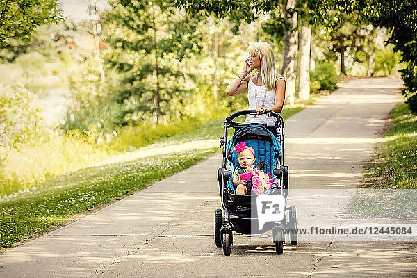A beautiful young mother taking her baby daughter out for a walk using a stroller in a park with a lake on a warm sunny day and talking on her cell phone  Edmonton  Alberta  Canada
