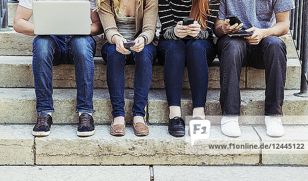 Vier Studenten,  die in einer Reihe auf einer Stufe sitzen und ihre Technologie auf dem Universitätscampus nutzen,  wobei nur ihre Füße und Beine zu sehen sind,  Edmonton,  Alberta,  Kanada