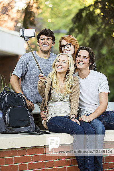 Eine Gruppe von vier Freunden sitzt auf einer Bank und macht ein Selbstporträt mit einem Smartphone und einem Selfie-Stick auf dem Universitätscampus in Edmonton,  Alberta,  Kanada