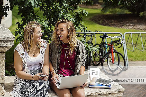 Zwei Studentinnen lachen und unterhalten sich  während sie draußen auf dem Campus ihre Technik benutzen  Edmonton  Alberta  Kanada