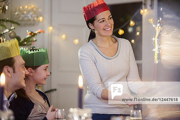 Glückliche Mutter in Papierkrone serviert Weihnachtspudding mit Feuerwerk bei Kerzenlicht am Tisch