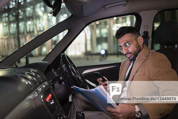 Businessman with smart phone reading paperwork in car at night