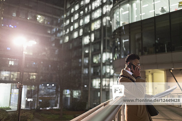 Businessman talking on smart phone,  reading paperwork on urban pedestrian bridge at night