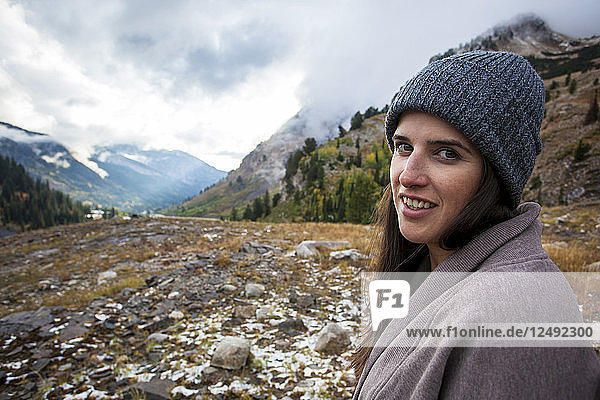 Eine Frau mit dunkelbraunem Haar steht vor dem Little Cottonwood Canyon und lächelt an einem kalten Herbsttag in die Kamera.