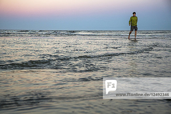 A man in shorts and a jacket walks through shallow water under a lavender sunset sky.