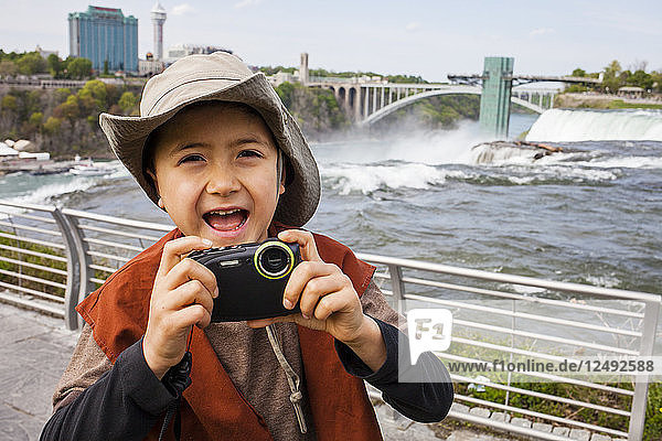 A 6 year old Japanese American boy is ready with his camera while exploring the overviews at Niagara Falls. He is standing in Niagara Falls  New York  USA  with views behind him of rainbow bridge and city skyline of Niagara Falls  Ontario  Canada.