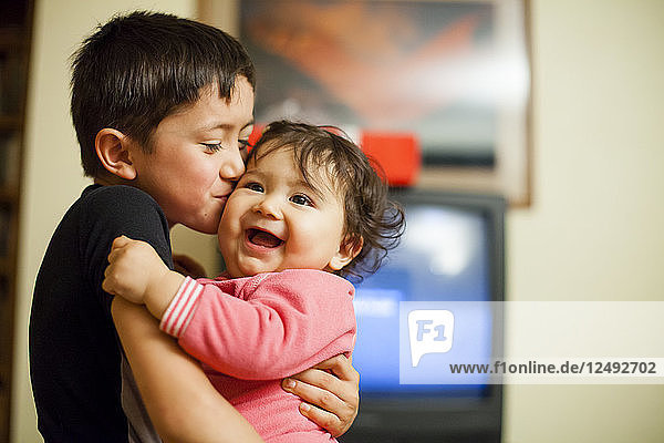 Ein einjähriges japanisch-amerikanisches Mädchen lacht  während es von seinem siebenjährigen Bruder im Wohnzimmer gehalten und geküsst wird.