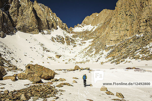 A 30 year-old man in mountaineering clothes walks up a snow-filled valley with jagged granite walls.
