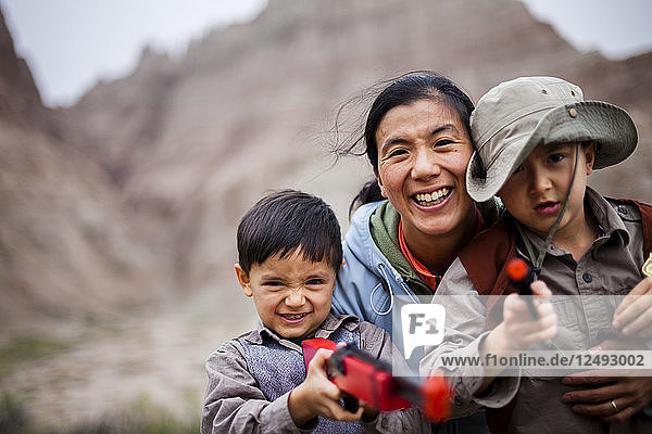 Eine japanisch-amerikanische Frau und Mutter hockt mit ihrem 4 Jahre alten japanisch-amerikanischen Jungen und ihrem 6 Jahre alten amerikanischen Jungen  die jeweils mit Spielzeugpistolen  einer 6-Schuss-Pistole und einem Gewehr ausgerüstet sind  inmitten von erodierenden Felsformationen (Zinnen und Türme) im Badlands National Park  South Dakota.
