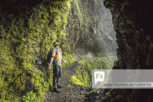 A Man Standing At The Opening Of A Tunnel Along A Lush Green Moss-lined Trail With A Waterfall Falling From Above