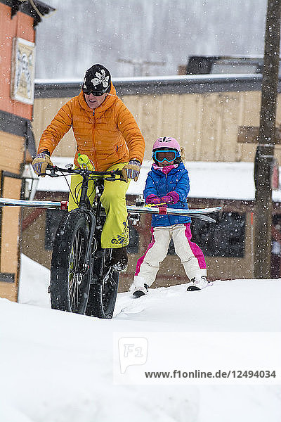 Eine Mutter zieht ihre Tochter mit Skiern auf einem Fahrrad mit großen Reifen durch die verschneiten Straßen von Silverton  Colorado.