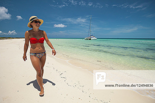 Eine junge Frau mit Bikini und Sonnenhut spaziert im Urlaub auf Kuba über einen Sandstrand.