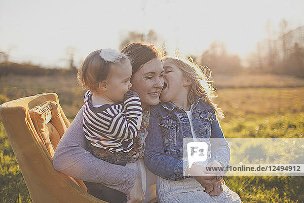 Eine Mutter hält ihre beiden Töchter  während die eine sie auf die Wange küsst.