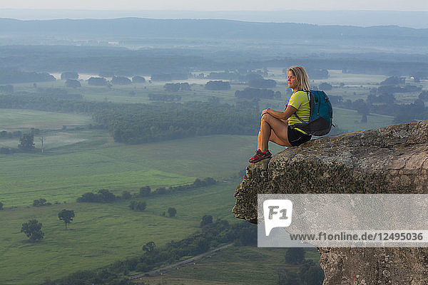 Frau sitzt auf einem Sandsteinüberhang und betrachtet den Sonnenaufgang vom Gipfel des Petit-Jean Mountain über dem Arkansas River Valley